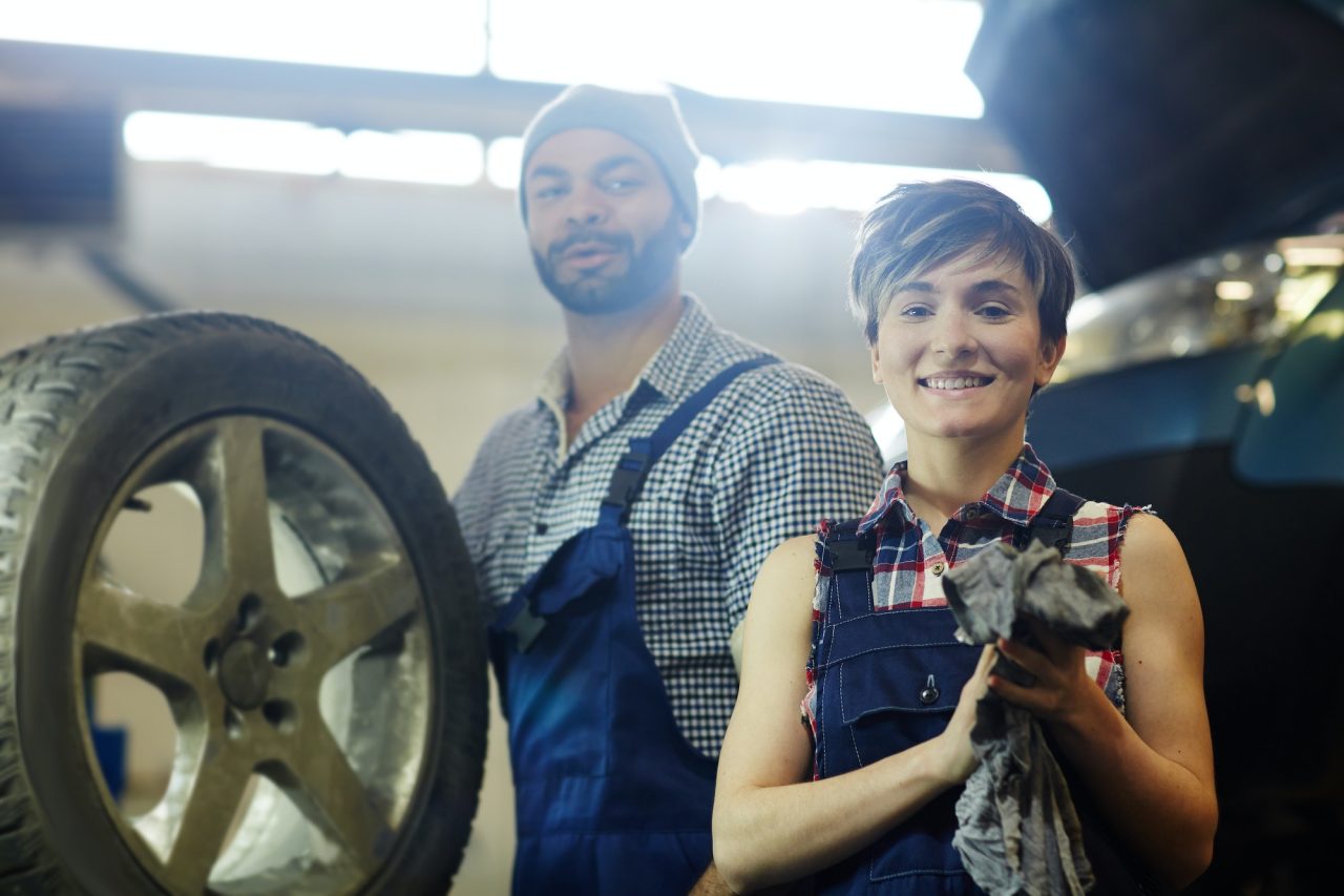 two mechanics smiling next to a car wheel