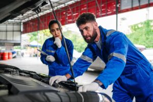 Two auto mechanics in blue and white coveralls inspect and perform maintenance on a car engine.