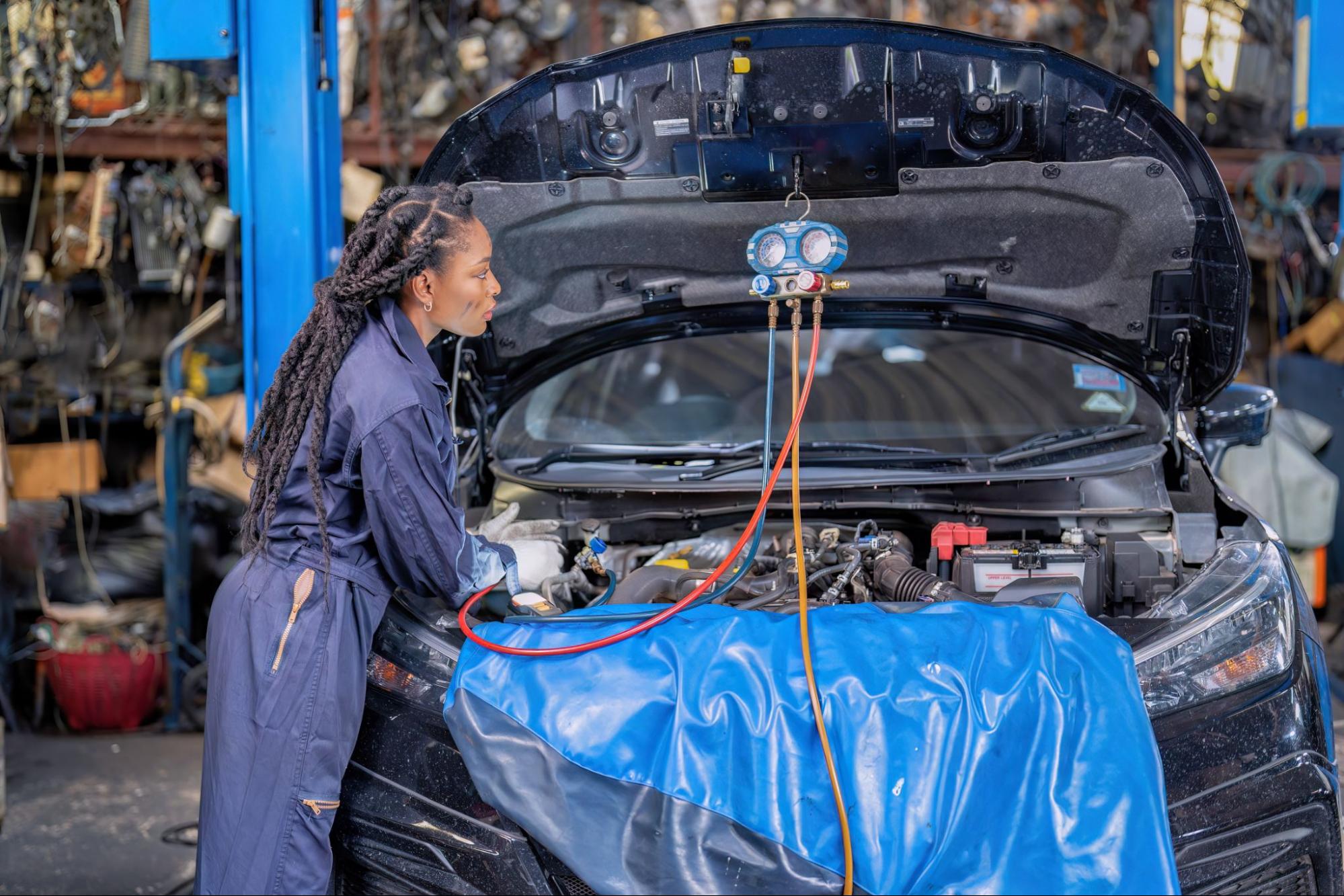 A female mechanic in coveralls checks a car engine with a hanging diagnostic meter. A blue tarp covers part of the engine.